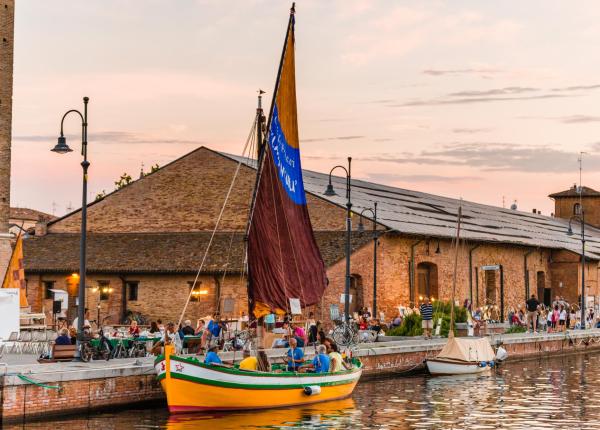 Barca a vela gialla sul canale, edifici storici e persone lungo la riva.